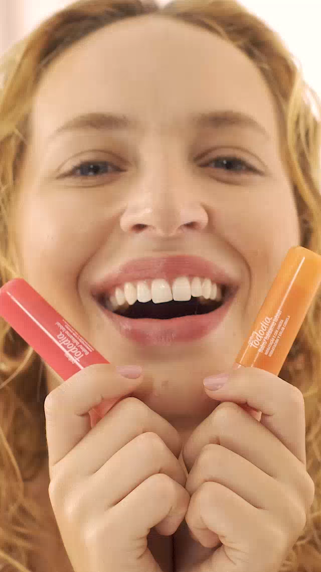 Uma mulher loira, com brincos de argola, sorri para a câmera enquanto segura dois balm labiais de Tododia em formato de bastão. Um é rosa e o outro é laranja. Ela pisca, faz um bico e aproxima os produtos dos lábios, como se estivesse aplicando. Depois, volta a sorrir para a câmera.