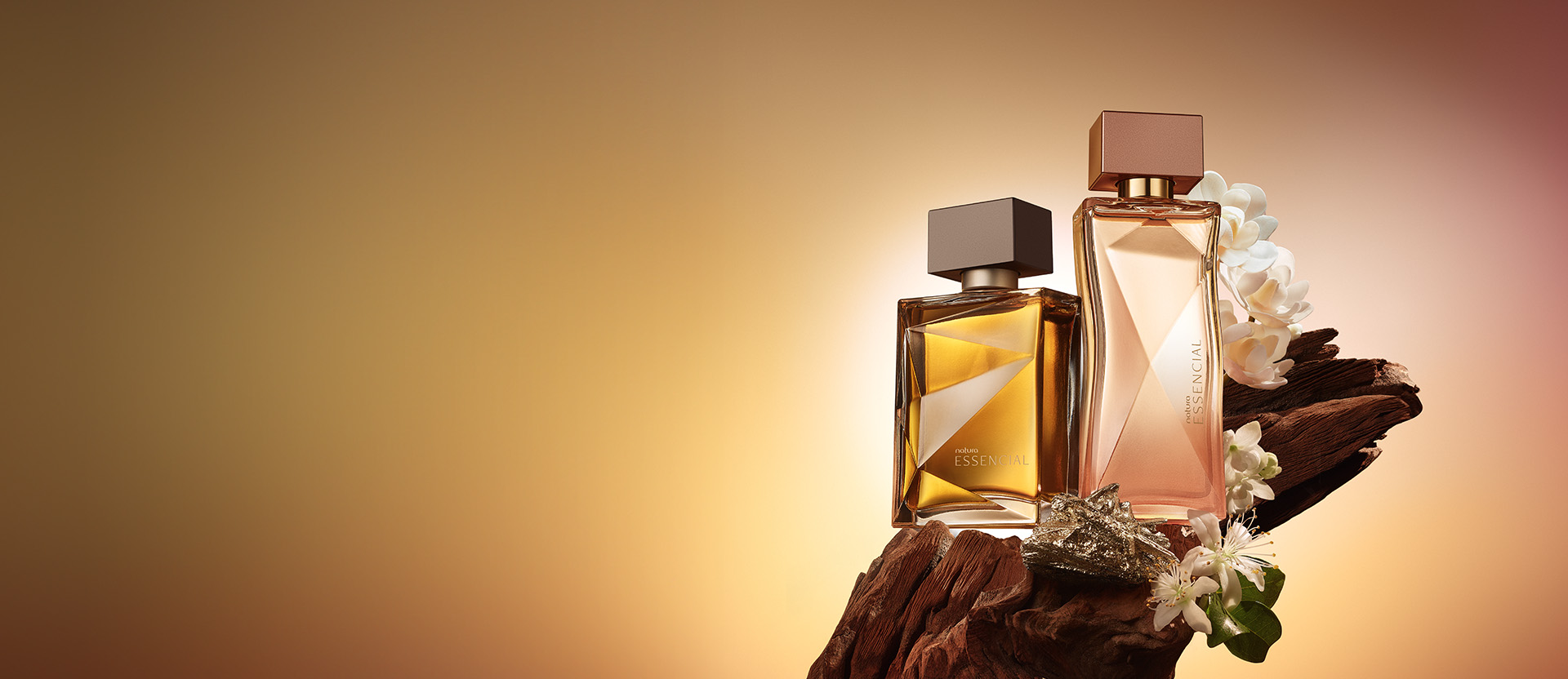 Dois frascos de perfume Natura Essencial, um em tom âmbar e outro em tom pêssego/rosado, posicionados sobre um pedaço de madeira rústica com flores brancas e um elemento metálico prateado. O fundo é um gradiente quente, em tons de amarelo-ouro e marrom. A embalagem âmbar é o 'Natura Essencial'."