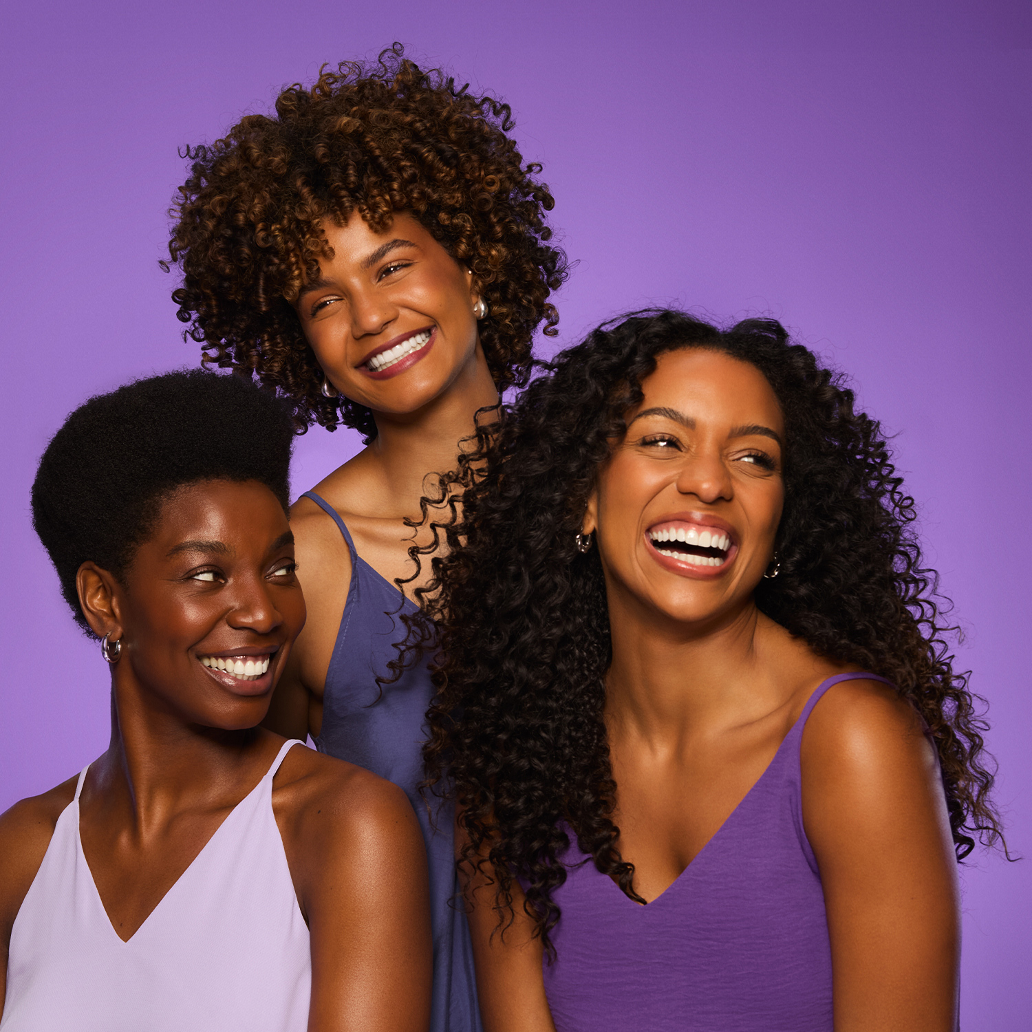 3 mulheres jovens sorriem desviando o olhar da câmera. Elas vestem brulas lilás, estão em frente a um fundo roxo e tem cabelos crespos volumosos, definidos e brilhantes.