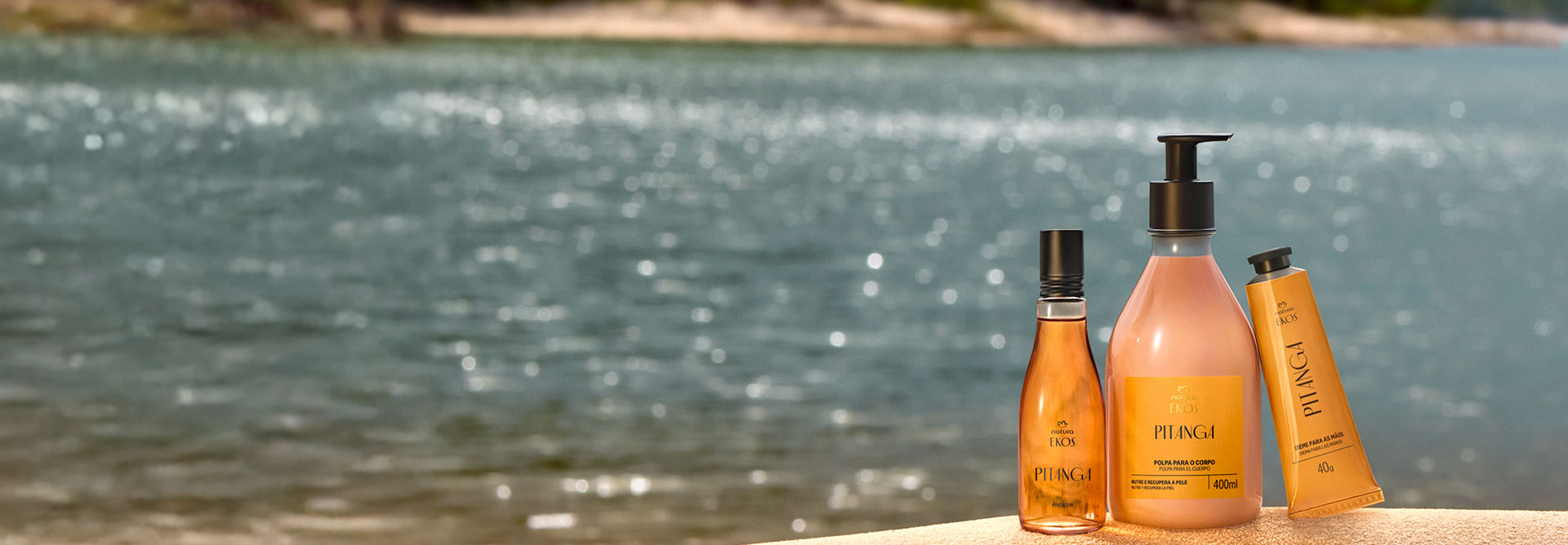 Três produtos Natura Ekos Pitanga estão sobre uma superfície bege: Desodorante Colônia Frescor, Creme Hidratante o Corpo e Creme para Mãos. Ao fundo, rio azul com brilho suave e margem desfocada.
