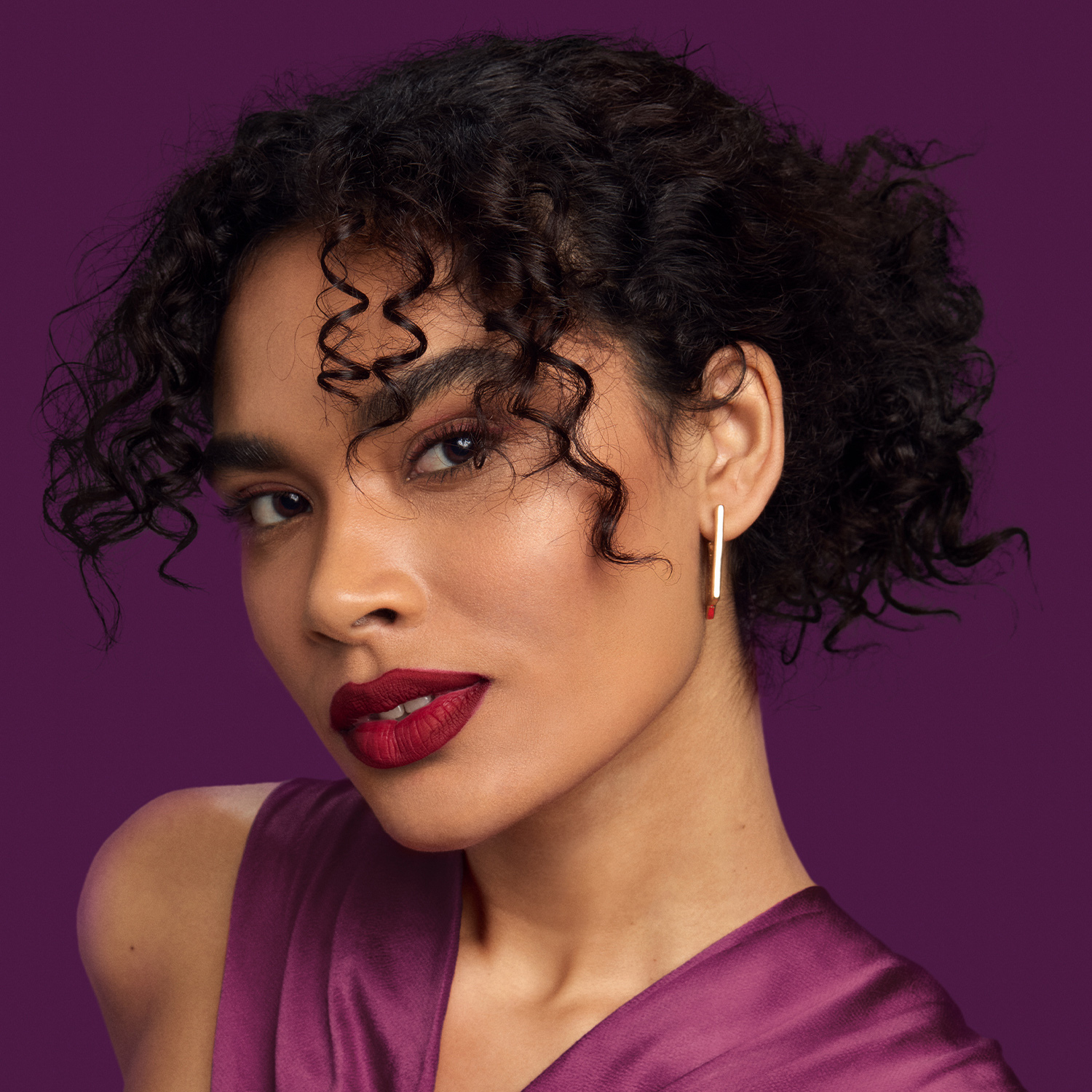 Mulher de cabelo crespo preso em um coque despojado olha em direção à câmera. O fundo é roxo. Ela veste uma blusa de cetim roxa e usa  batom vermelho. 