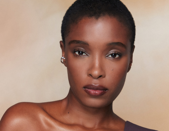 Close de rosto de mulher com pele negra, cabelo curto e maquiagem discreta; ombro nu visível. Fundo bege suave, olhar direto à câmera.