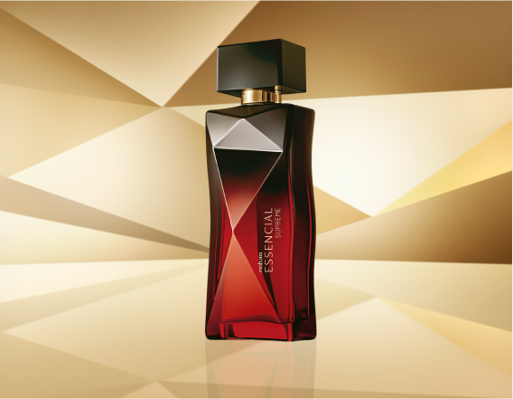 Frasco de Essencial Supreme Feminino, em tons de vermelho e preto com tampa preta, aparece no centro da tela sobre fundo multifacetado dourado, fazendo alusão ao design do frasco de Essencial.