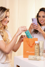 Imagem mostra mulher loira de pele clara colocando itens Natura Tododia para Cabelos em uma sacola laranja da Natura. Uma mulher morena de pele clara e cabelos cacheados filma a cena com o celular.