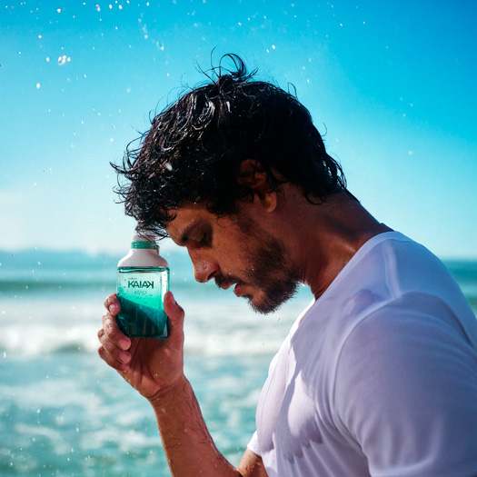Homem de cabelos escuros e camiseta branca na praia com mar e céu azul ao fundo. Ele segura junto à têmpora frasco verde com gotas de água na embalagem da fragrância Kaiak Aero.