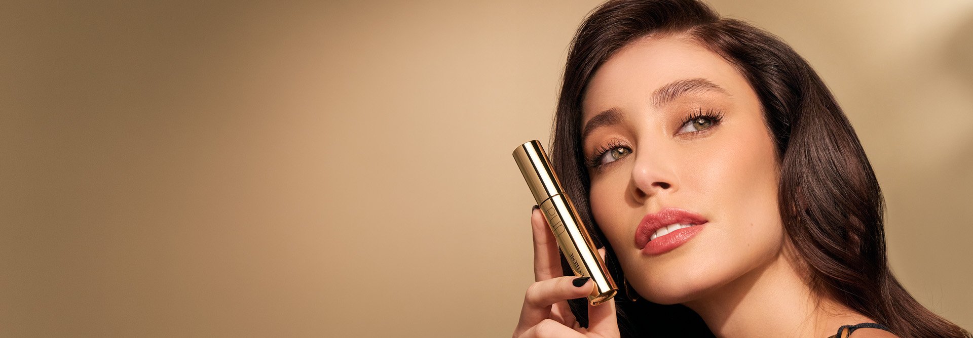 Mulher branca de cabelos escuros e longos e cílios curvados segura a Máscara de Cílios Extreme Multibenefícios Natura Una nas mãos. Fundo bege-claro.