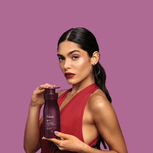 Mulher de cabelos castanhos e presos usando vestido vermelho segura um produto Natura Tododia, em fundo lilás.
