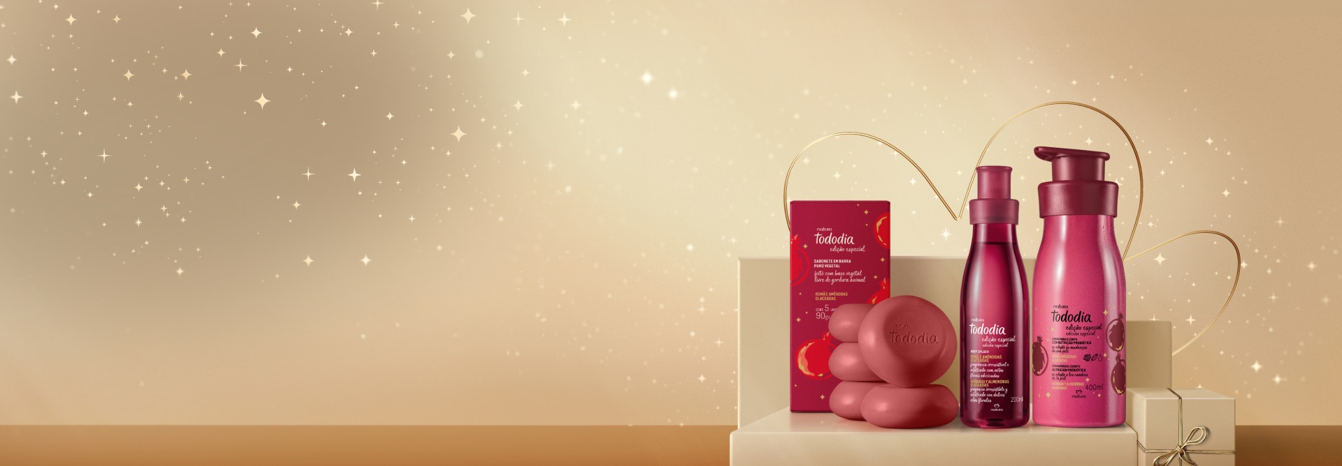 Presente Natura Tododia Romã e Amêndoas Glaceadas, com Body Splash, Creme Desodorante Corporal e Caixa de Sabonetes, em fundo claro com estrelas e flor símbolo da Natura. Embalagens vermelhas.