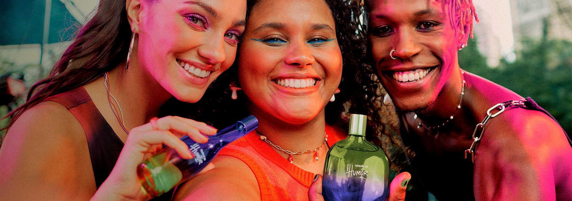 Selfie com duas mulheres e um homem sorrindo, segurando e interagindo com os frascos de Natura Humor Festivais.