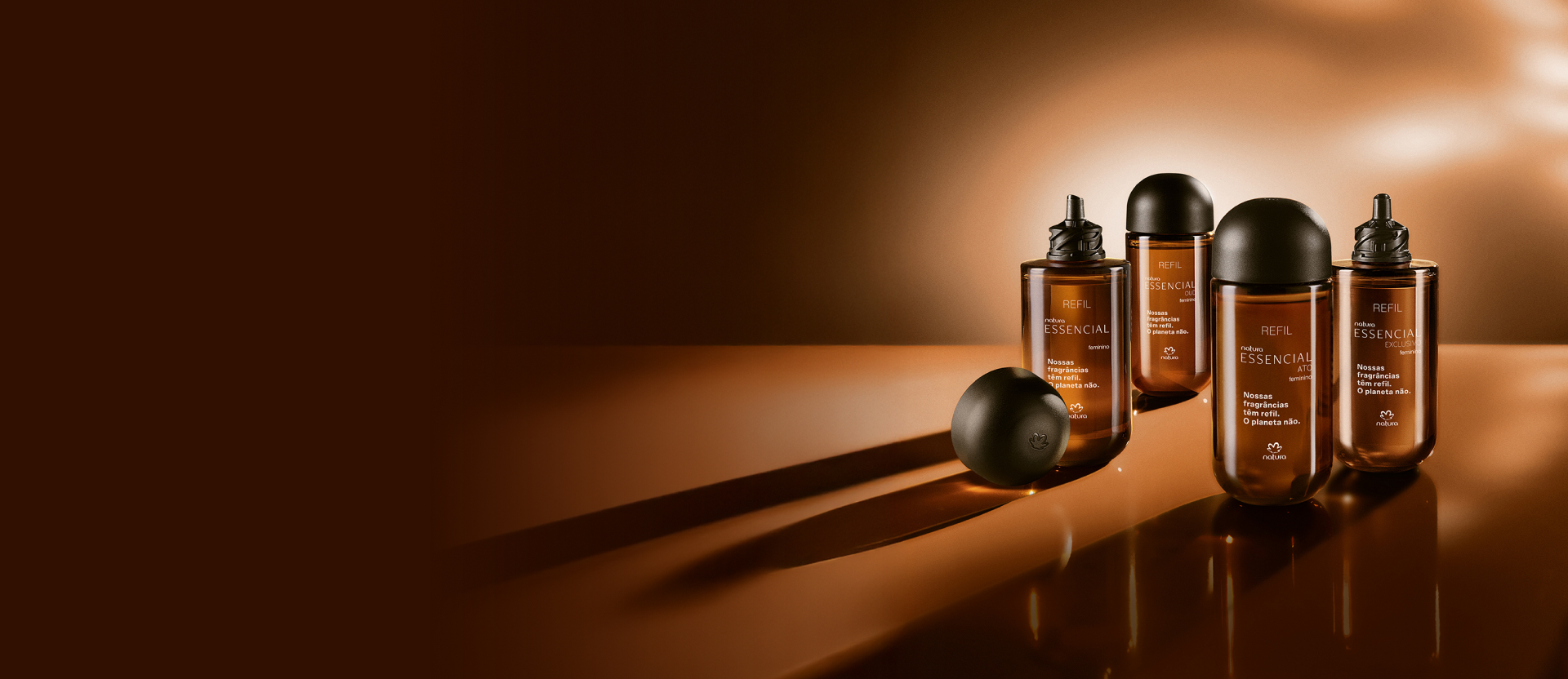 Frascos de refis de vidro âmbar e tampas arredondadas das fragrâncias femininas Essencial, Essencial Oud, Essencial Ato e Essencial Exclusivo aparecem iluminados sobre fundo em tons de marrom.