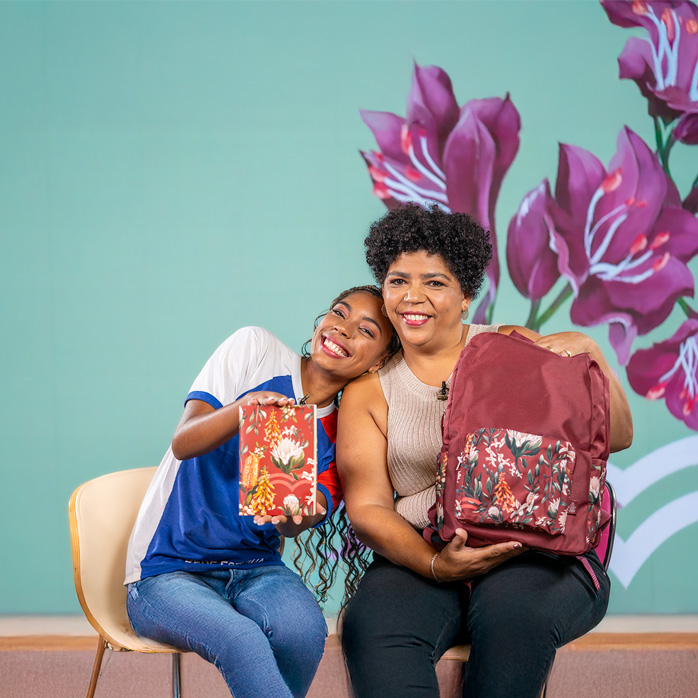 Duas mulheres sorridentes de pele escura em um fundo verde-claro com desenho floral roxo. A mulher mais jovem, à esquerda, com camiseta branca e azul, apoia a cabeça no ombro da mais velha e segura um caderno estampado. A mulher mais velha, à direita, veste uma blusa bege e segura uma mochila vinho com estampa floral.
