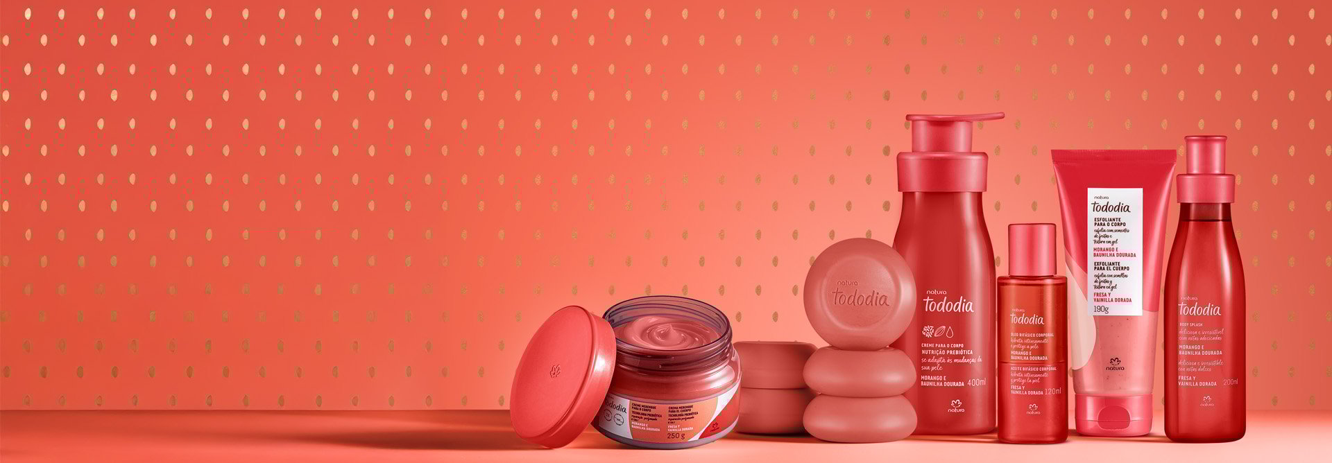 Produtos da linha Natura Tododia "Morango e Baunilha Dourada" organizados sobre uma superfície vermelha. A imagem mostra um pote de creme aberto, sabonetes em barra empilhados, hidratante corporal com válvula pump, óleo bifásico, esfoliante em bisnaga e um body splash. O fundo é rosa com pequenos detalhes dourados.