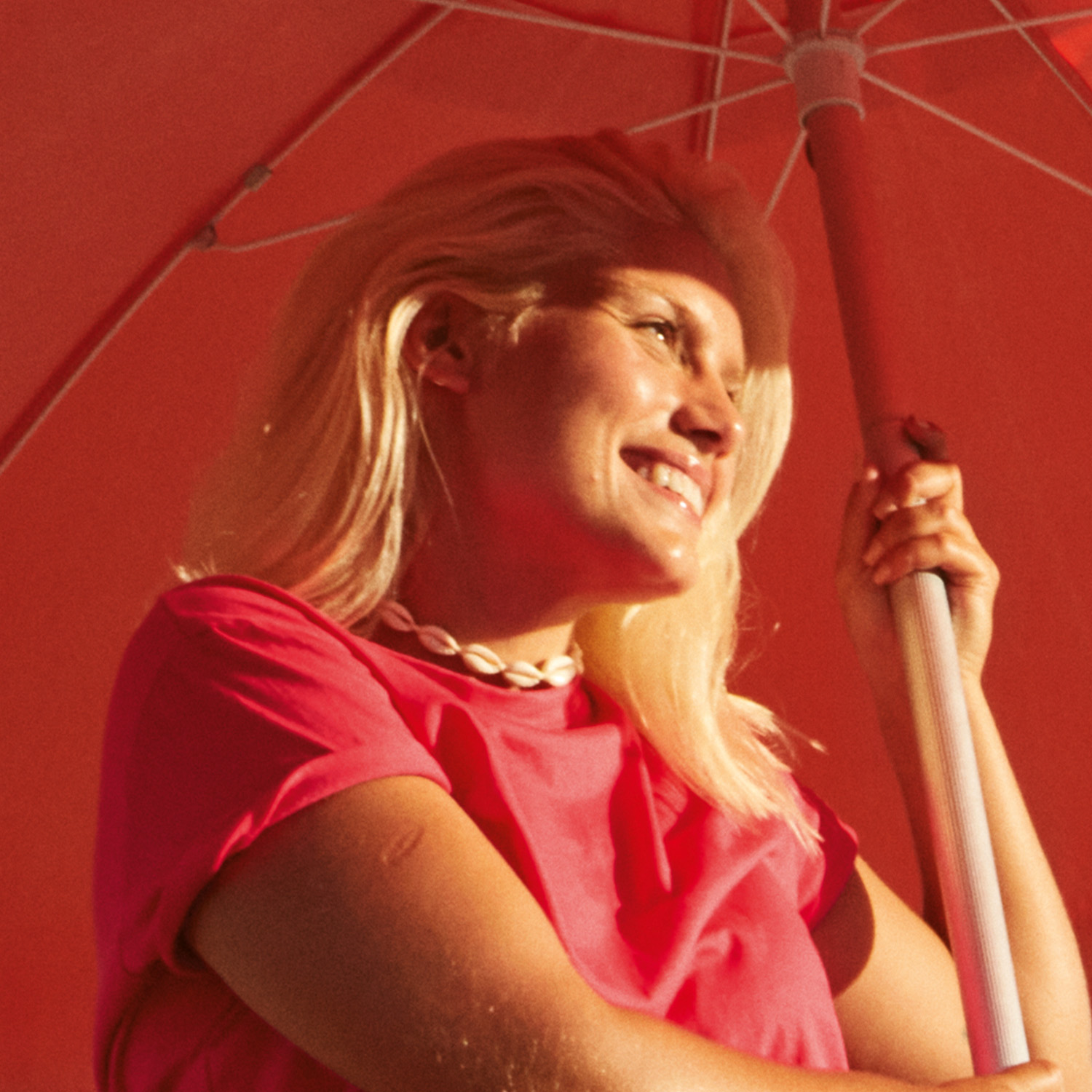 Mulher jovem de cabelo platinado, usando camiseta rosa e colar de conchas, segura um guarda-sol rosa e olha para o horizonte.