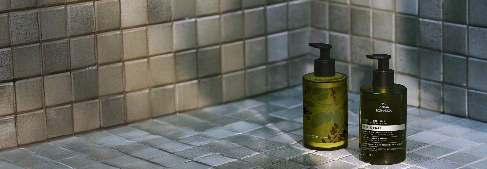Fundo e superfície com ladrilhos cinzas. Em destaque no centro, Sabonete Líquido e Hidratante para Mãos da linha Natura Bothânica com frascos em cor verde e tampa preta.