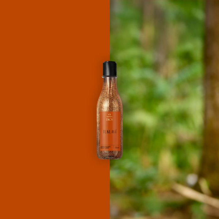 Frasco transparente do Sabonete Líquido Esfoliante Corporal Ekos Tukumã com tampa preta e rótulo laranja ao centro da imagem. Fundo dividido entre laranja sólido e floresta desfocada em tons de verde.