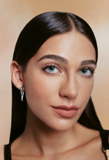 Rosto de mulher branca com maquiagem natural: pele lisa, sobrancelhas definidas, cílios longos, lábios rosados; cabelo castanho-escuro liso penteado para trás. Fundo bege suave e uniforme.