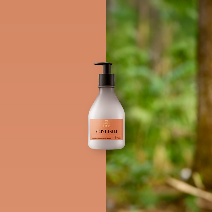 Frasco do Ekos Castanha Hidratante para Mãos 250 ml ao centro, com tampa preta. Ao lado, castanhas abertas sobre fundo bege e folhas verdes desfocadas à esquerda.