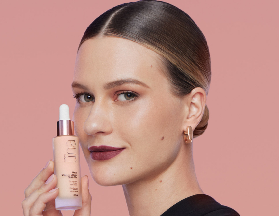 Foto de mulher com cabelos presos e loiros segurando um produto Natura Una próximo ao rosto, em fundo rosa-claro.