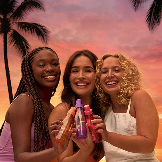 Três mulheres sorridentes, de diferentes etnias e tipos de cabelo, estão em pé, lado a lado, ao ar livre, segurando e brindando com pequenos frascos coloridos (laranja, roxo e rosa/vermelho). O fundo é um céu de pôr do sol vibrante com tons de laranja, rosa e amarelo, e silhuetas de palmeiras.