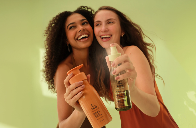 Foto de duas mulheres sorrindo sobre fundo verde claro, segurando produtos Natura Tododia, uma com hidratante corporal e outra com body splash.