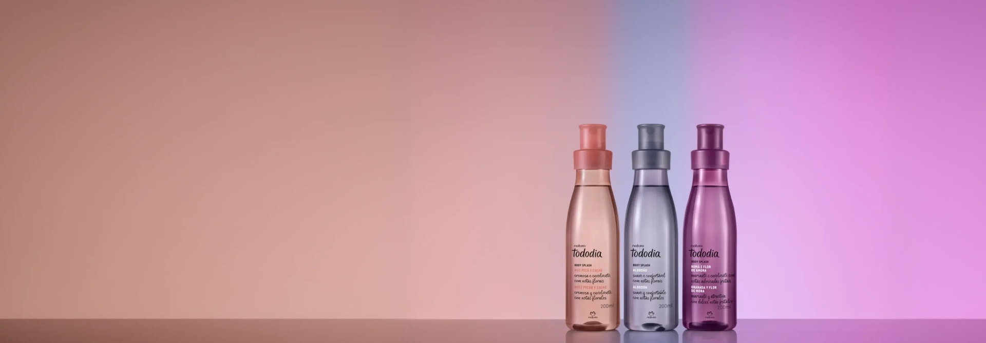 Três frascos translúcidos coloridos de body splash Tododia: 1 Noz Pecã e Cacau, 1 Algodão e 1 Romã e Flor de Amora. Fundo degradê com tons de rosa, lilás e roxo, nas mesmas cores dos frascos.