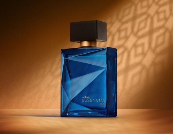 Deo Parfum Essencial Oud Masculino em frasco facetado azul com tampa preta. Está em fundo cor areia com sombra geométrica projetada.