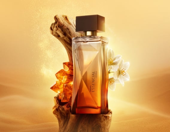 Deo Parfum Essencial Mirra Feminino em frasco degradê âmbar com tampa preta. Está em fundo areia sobre um tronco de madeira escura com flores brancas e pedras âmbar ao redor.