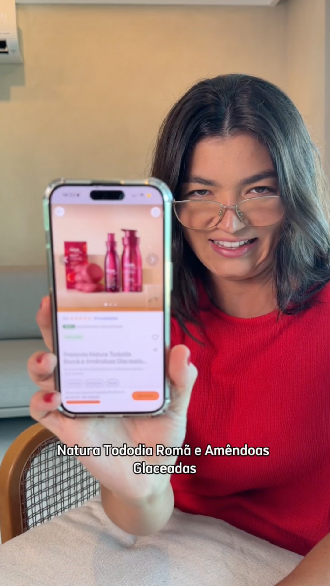  Lara, uma jovem mulher branca de cabelos longos e escuros, usa óculos e uma blusa vermelha. No início, ela aparece reagindo a um vídeo no celular, rindo e comentando. Depois, ela é vista decorando uma parede com folhagens e festão dourado, usando um gorro de Papai Noel. Ela canta uma paródia de música natalina e mostra na tela do celular o kit Natura Tododia Romã e Amêndoas Glaceadas. O vídeo tem um tom caseiro e bem-humorado, terminando com Lara sorrindo para a câmera.