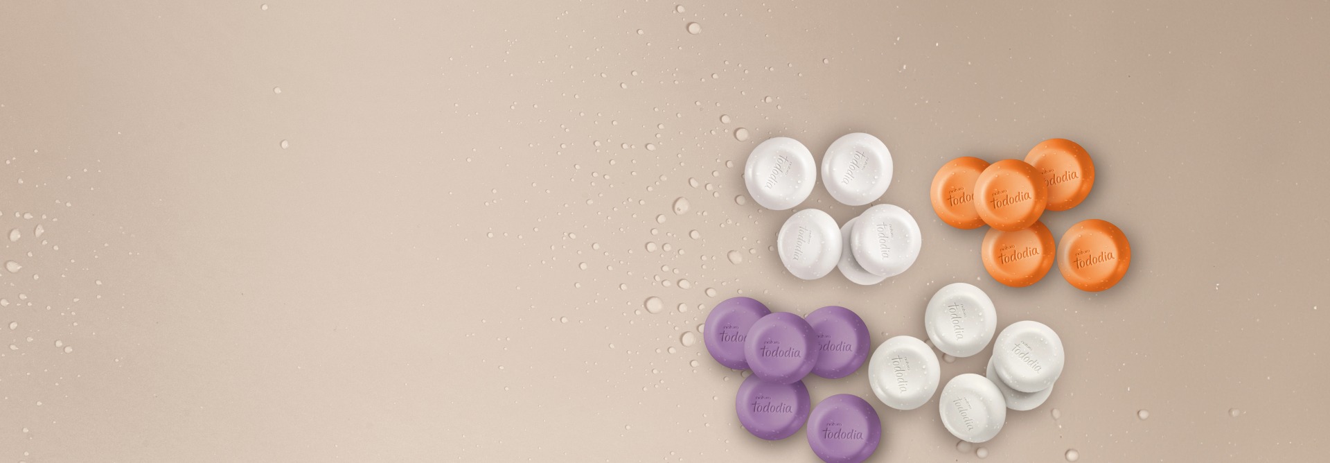 Sabonetes em Barra Tododia espalhados sobre um fundo bege com gotas de água. São 20 barras no total, com formato redondo, a marca Natura Tododia esculpida e nas cores branco, laranja e roxo.