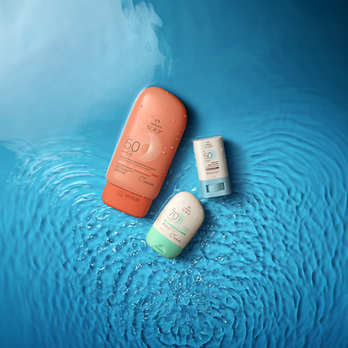 rês itens na diagonal em fundo azul aquoso: embalagem laranja de Protetor Solar Corporal, stick branco com azul de Protetor Solar Facial e embalagem branca com tampa verde de Protetor Solar Facial.