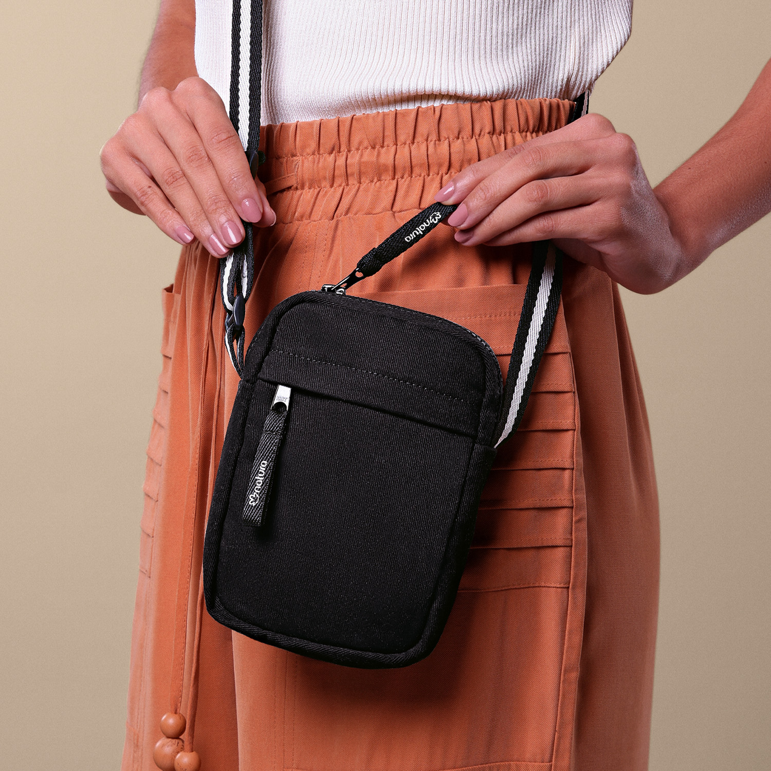 Foto com zoom. Mulher de calça laranja abre a bolsa transversal preta de Kaiak. A alça da bolsa está apoiada no ombro e tem detalhe de listras pretas e brancas. 