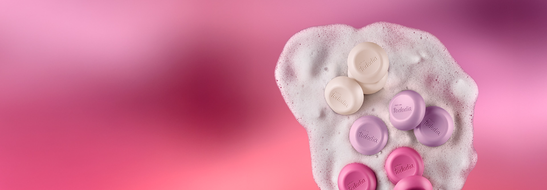 Diversos sabonetes nas cores branco, lilás e rosa sobre espuma cremosa branca. Fundo em tons de rosa.