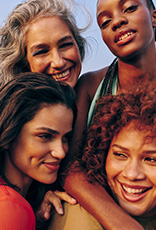 Quatro mulheres sorrindo em um abraço coletivo. A mulher no canto superior esquerdo tem cabelos grisalhos longos. A mulher na parte inferior esquerda tem pele escura e cabelos trançados. A mulher no centro tem cabelos cacheados volumosos e castanho-avermelhados. A mulher no canto superior direito tem cabelos escuros e ondulados.