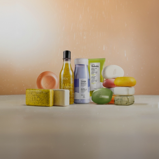 Gotas de água caem sobre sabonetes em barra, sabonetes líquidos e esfoliantes corporais Natura Ekos e Natura Tododia em destaque no centro da imagem sobre fundo rosado.