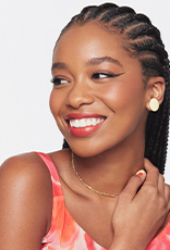 Mulher negra de cabelo escuro trançado sorri olhando para o lado. Ela usa batom vermelho, delineado preto, brinco e colar dourados e uma blusa de alça rosa. Fundo branco.