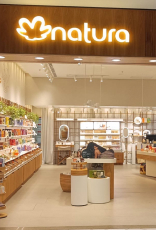 Imagem mostra uma loja física da Natura. Na entrada, logo da Natura iluminado sobre fachada de madeira e vitrine decorada. Dentro da loja, produtos organizados por categorias.