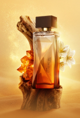Deo Parfum Essencial Mirra Feminino em frasco degradê âmbar com tampa preta. Está em fundo areia sobre um tronco de madeira escura com flores brancas e pedras âmbar ao redor.