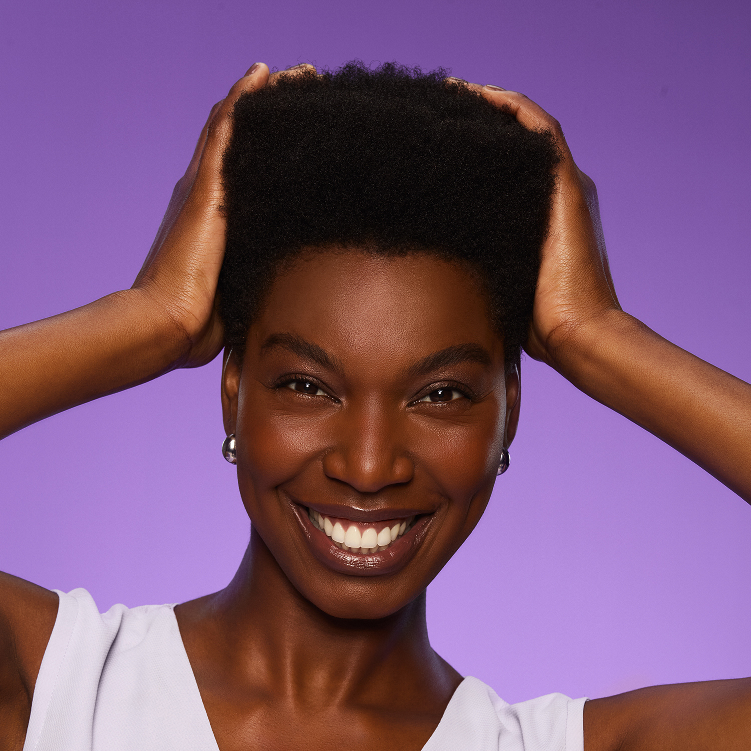 Mulher negra com sorriso largo e gloss nos lábios toca com orgulho em seu cabelo crespo preto. O cabelo é curto nas laterais e volumoso no topo, com fios de aparência leve e com brilho.