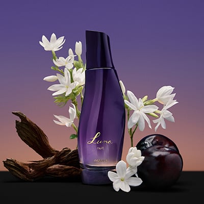 Frasco roxo-escuro do Desodorante Colônia Luna Nuit rodeado por flores brancas, galhos de madeira escura e ameixa. Fundo degradê roxo e laranja.