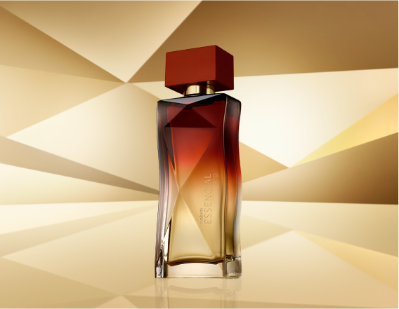 Frasco de Essencial Ato Feminino, nos tons de vermelho-queimado e dourado, aparece em destaque no centro da tela sobre fundo multifacetado dourado, fazendo alusão ao design do frasco de Essencial.