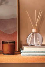 Vela Perfumada e Difusor Natura 861 Guaiaco Pataqueira aparecem em destaque sobre prateleira de madeira. O Difusor repousa sobre dois livros e a Vela Perfumada destaca-se à frente de tela abstrata.