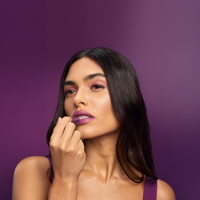 Mulher de cabelos soltos e lisos aplicando o Balm Labial Natura Tododia em fundo roxo.
