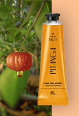 Bisnaga amarela do Creme Hidratante para as Mãos Ekos Pitanga à direita, ao lado de uma pitanga vermelha com gota de extrato, sobre fundo de folhas verdes e laranja suave.