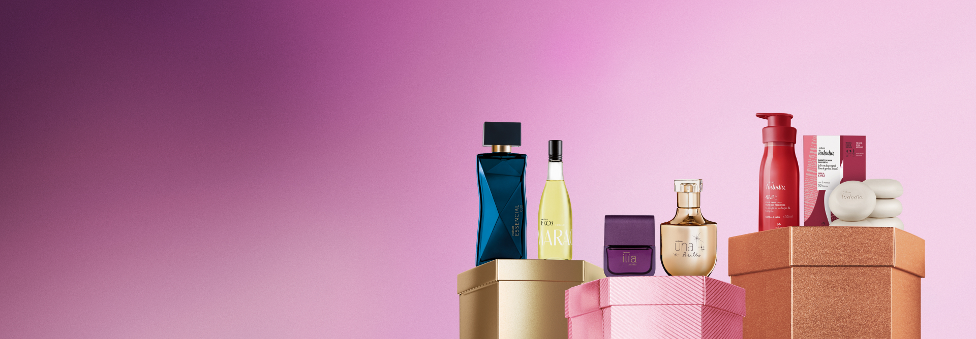 Embalagens sobre caixas brilhantes dourada, rosa e marrom, em fundo rosa: frasco de frescor Ekos Maracujá, fragrâncias Essencial Oud, Ilía Secreto e Una Brilho, e loção e sabonetes com caixa Cereja e Avelã.