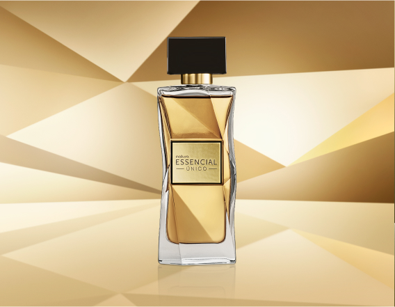 Frasco de Essencial Oud Feminino, em tom de azul-escuro e tampa preta, aparece em destaque no centro da tela sobre fundo multifacetado dourado, fazendo alusão ao design do frasco de Essencial.