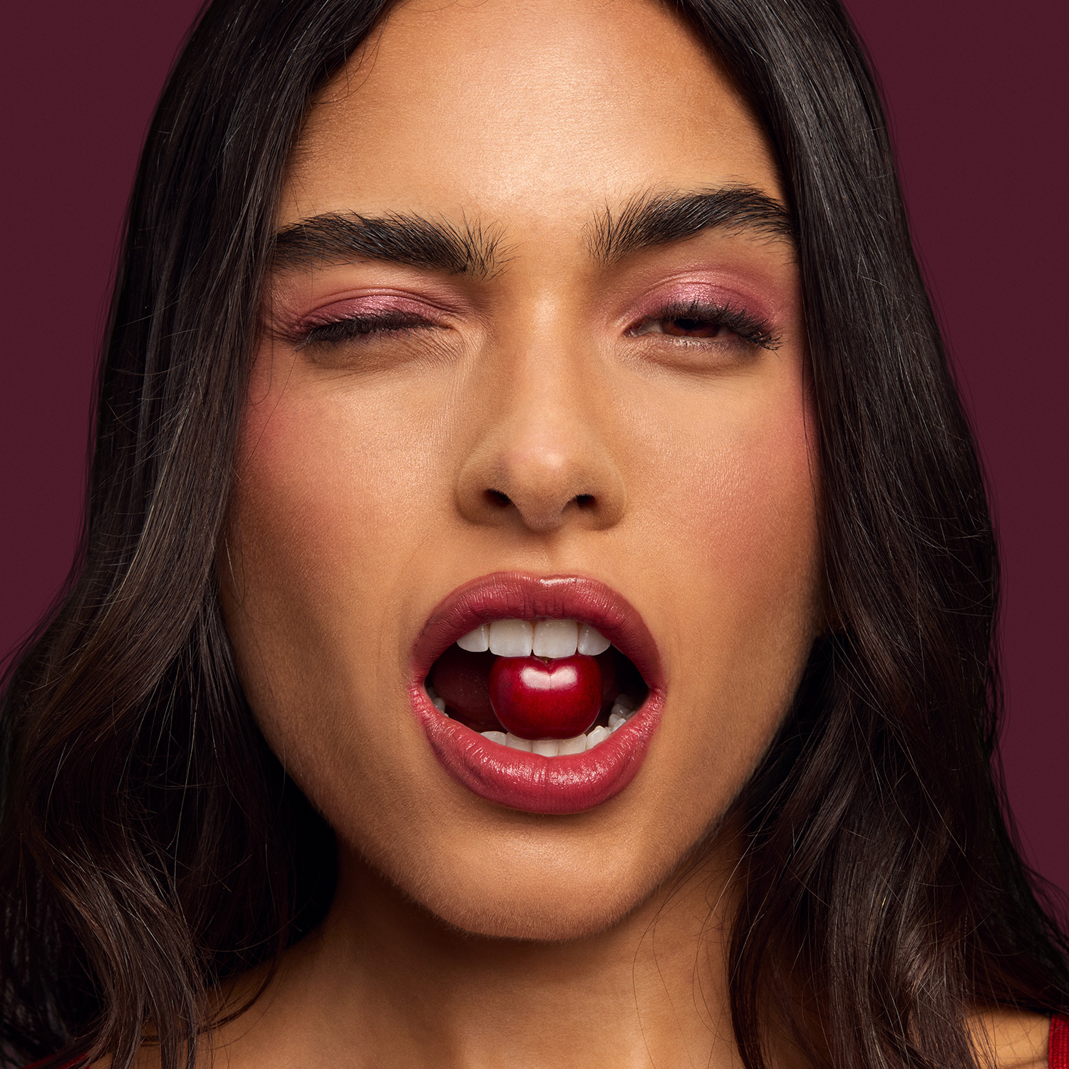 Mulher jovem de cabelo castanho ondulado pisca com um dos olhos e segura entre os dentes uma cereja. Ela usa maquiagem iluminada rosa, gloss nos lábios e blusa vermelha. Fundo roxo. 