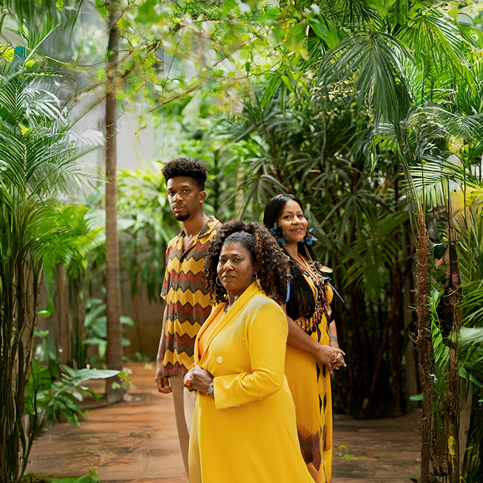 Três pessoas: uma mulher negra à frente, uma mulher indígena à direita e homem negro à esquerda, em ambiente com plantas amazônicas diversas.