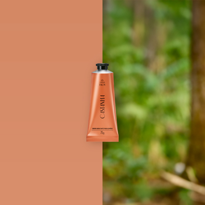 Bisnaga cobre do Creme Hidratante para Mãos Ekos Castanha 75 g ao centro, com castanhas abertas ao lado direito em fundo bege e folhas verdes desfocadas à esquerda.