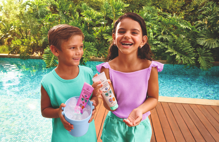 Duas crianças sorrindo segurando produtos Natura, em frente a uma piscina com jardim ao fundo.