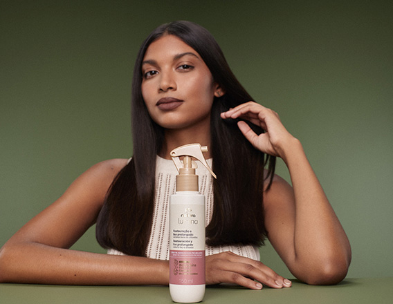 Em fundo verde, produto Natura Lumina à frente de mulher de cabelos longos e lisos.